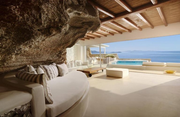 Suite con roca natural integrada, sofá blanco y piscina privada con vistas al mar en Cavo Tagoo Mykonos