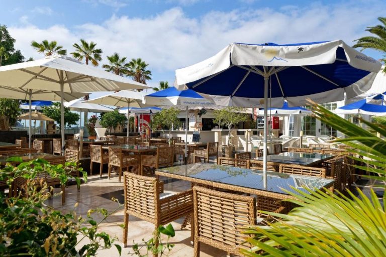 Terraza exterior del Aparthotel Maracaibo con mesas, sillas de ratán y sombrillas azules y blancas