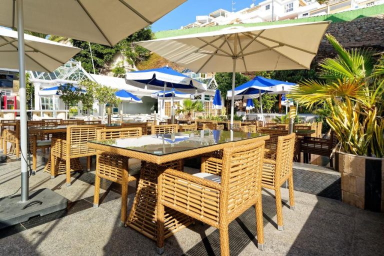 Terraza exterior con mesas y sillas de ratán del Aparthotel Maracaibo con sombrillas azules y blancas