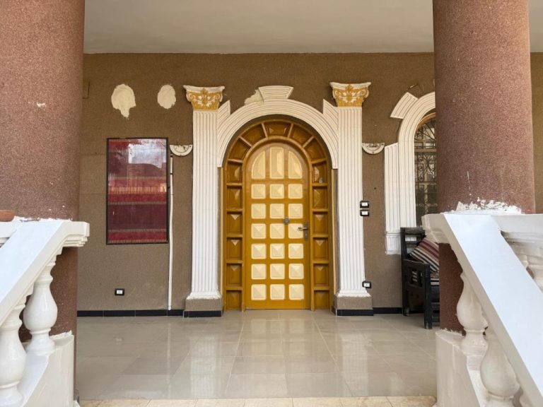 Entrada principal con puerta arqueada dorada y columnas blancas clásicas del Ramla Palace House