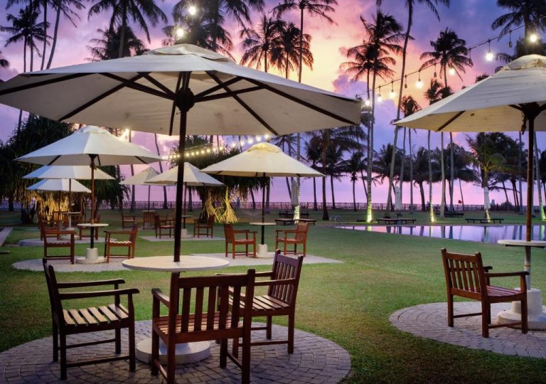 Terraza exterior con sombrillas y sillas de madera junto a la piscina al atardecer en The Blue Water Bawa Heritage Resort