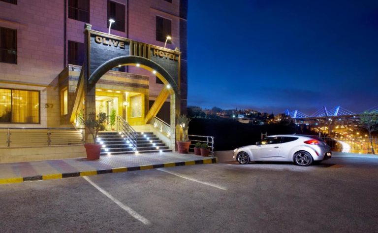 Entrada principal iluminada del Olive Hotel Amman de noche con aparcamiento y vistas a la ciudad