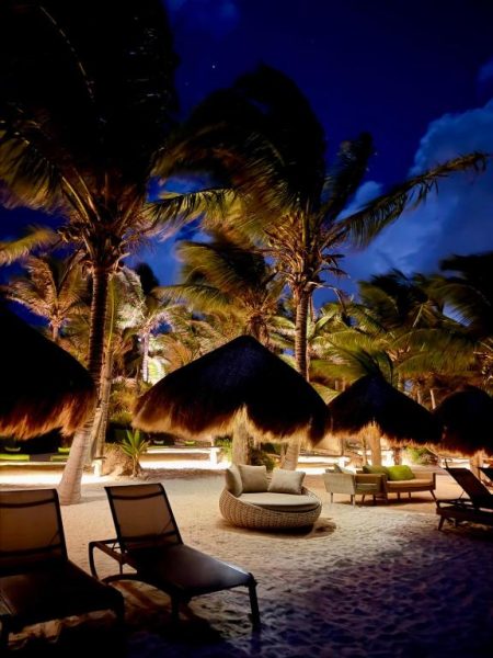 Terraza de playa iluminada por la noche con palmeras y camastros en Hotel Sueños Tulum