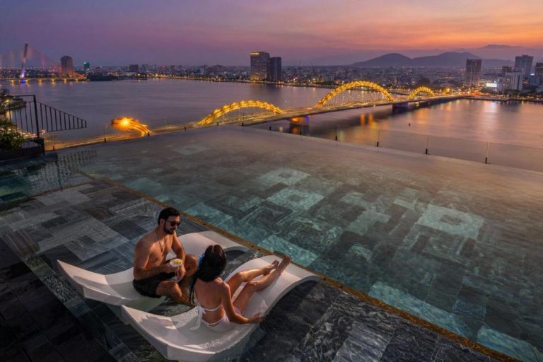 Pareja en piscina infinita del Caro Premium Danang Hotel con vistas al Dragon Bridge iluminado al atardecer