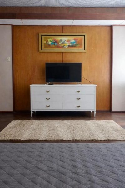 Habitación con balcón vista ciudad, cómoda blanca con televisor y alfombra beige en zona de descanso