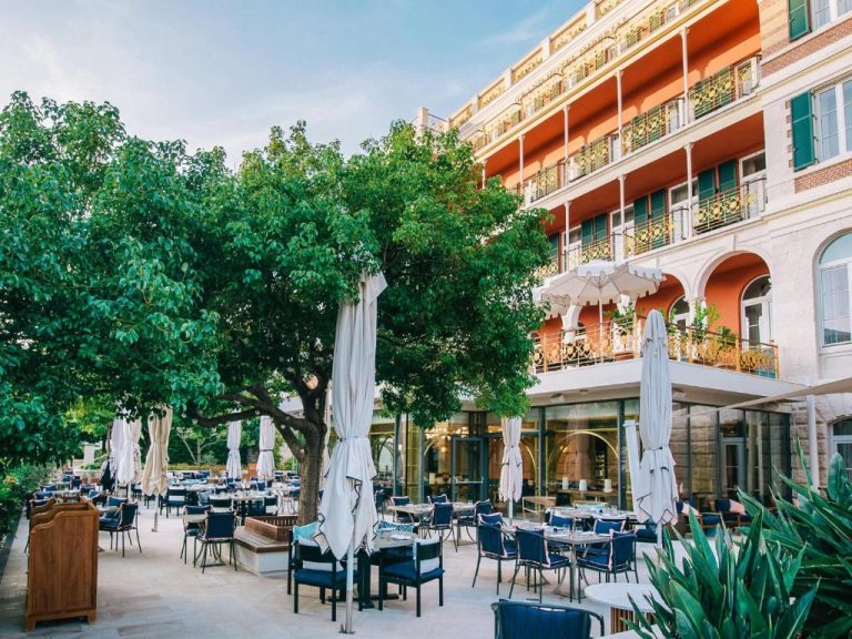 Terraza exterior del Hilton Imperial Dubrovnik con mesas, sillas azules y sombrillas blancas rodeada de árboles