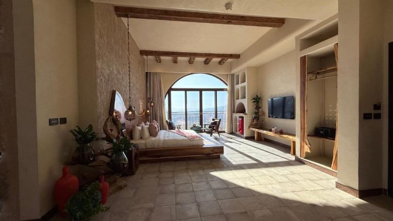Suite de lujo con cama de madera y ventanal arqueado con vistas panorámicas en Tal Rumman Boutique Hotel