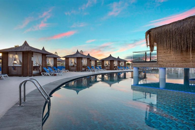 Piscina exterior con cabañas de madera y tumbonas azules al atardecer en Granada Luxury Belek