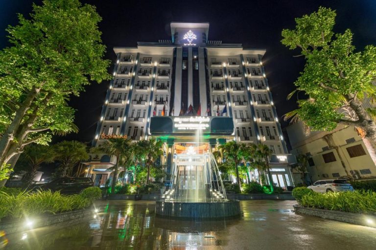 Fachada iluminada del Kampong Thom Royal Hotel con fuente ornamental y jardines tropicales de noche
