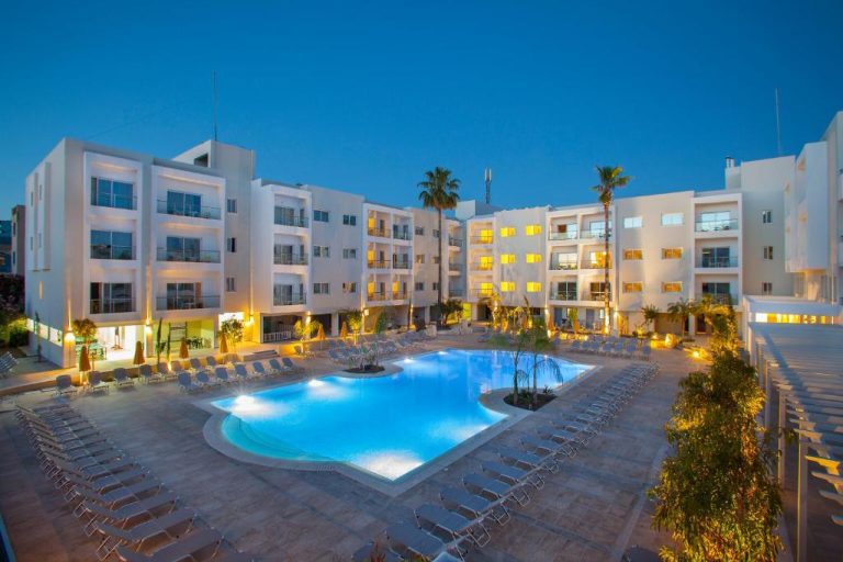 Piscina exterior iluminada del Mayfair Gardens rodeada de tumbonas y palmeras al atardecer