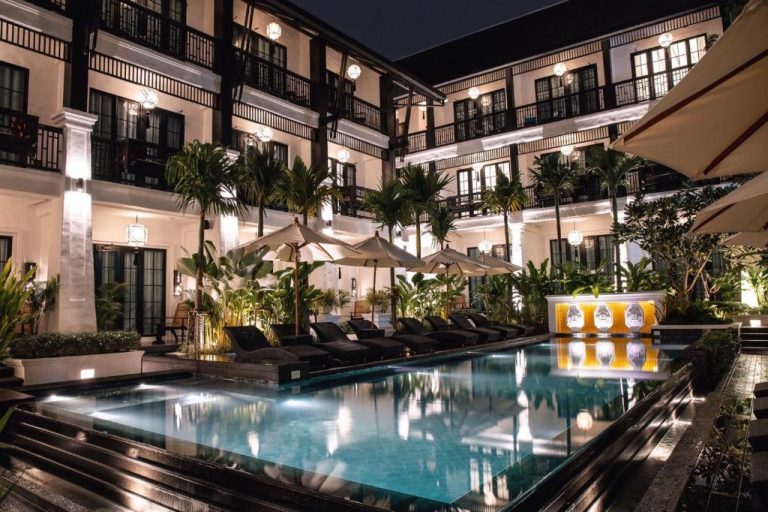 Piscina exterior iluminada de noche en el Hotel Aksara Heritage rodeada de palmeras y tumbonas