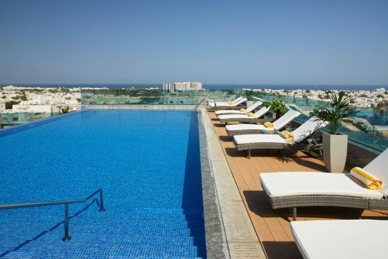 Piscina infinita en la azotea del Royal Tulip Muscat con tumbonas y vistas panorámicas a la ciudad y al mar