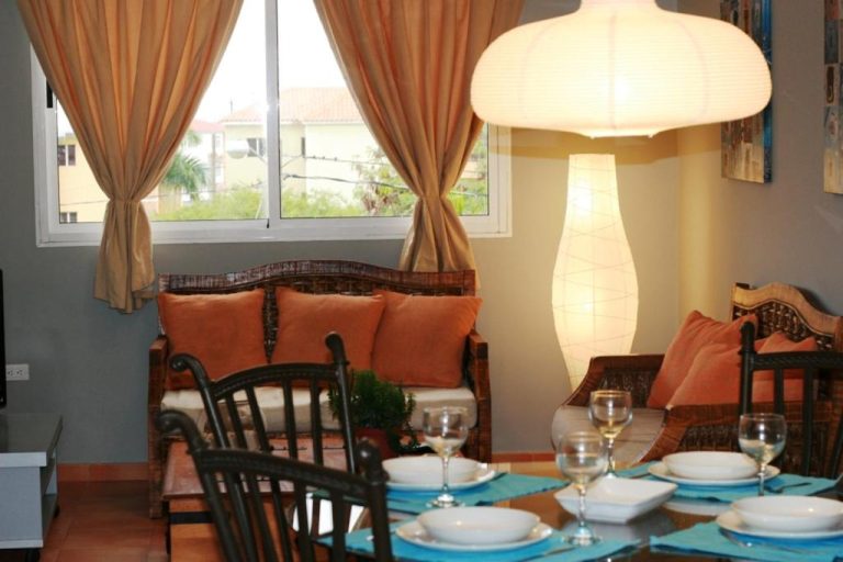 Salón comedor con mesa de cristal, sillas y sofás con cojines naranja en apartamento Bavaro Green