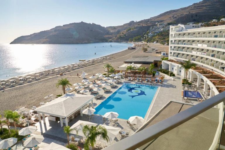 Vista aérea de la piscina exterior y playa del Lindos Bay Hotel con tumbonas y sombrillas en Rodas