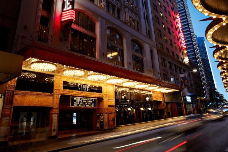 Fachada iluminada del QT Sydney junto al histórico State Theatre en el centro de Sídney de noche