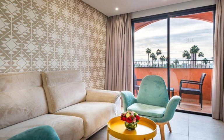 Salón con sofá y silla turquesa con terraza y vistas a palmeras en TUI BLUE Medina Gardens