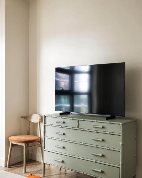 Habitación del ROOST Tampa con televisión sobre cómoda verde y silla de madera en apartamento moderno