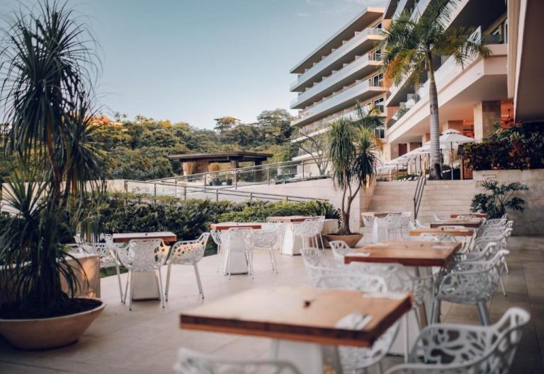 Terraza exterior del restaurante Wyndham Tamarindo con mesas de madera y sillas blancas rodeadas de vegetación tropical