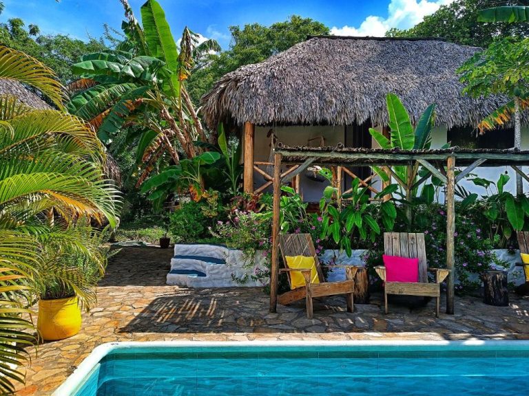 Piscina exterior con palapa y sillas de madera rodeada de vegetación tropical en Hotel Cabañas Puesta del Sol