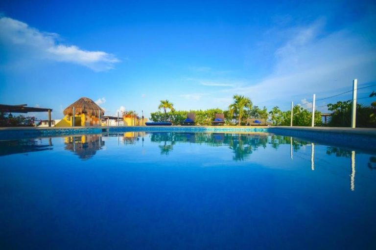 Piscina exterior de aguas cristalinas con palapa y palmeras del Hotel Encanto Riviera bajo cielo azul