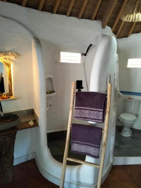 Baño rústico con ducha abierta y toallas moradas en el SHAYA Cottage, decoración tropical natural