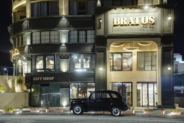 Fachada exterior nocturna del Bratus Hotel con coche clásico negro aparcado en la entrada iluminada
