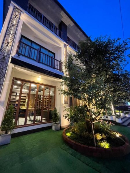 Fachada exterior iluminada del Baannalin Hometel con jardín y árbol decorativo en noche