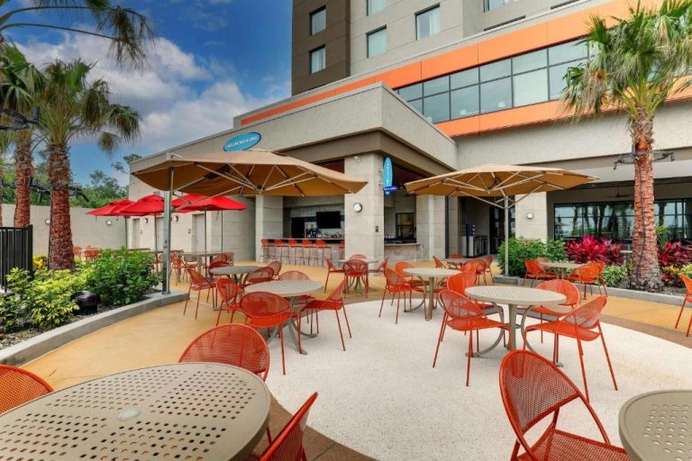 Terraza exterior con mesas y sillas del Drury Plaza Hotel Orlando Disney Springs Area con bar y palmeras