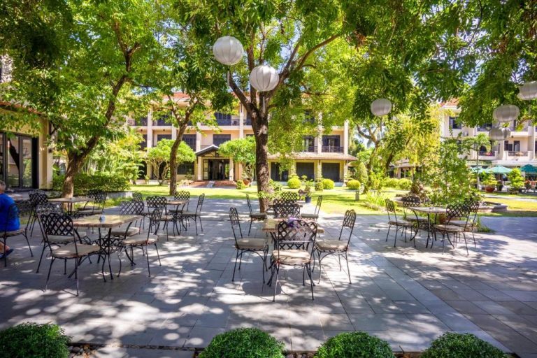 Terraza exterior con mesas y sillas de hierro forjado rodeada de jardines en Hoi An Historic Hotel