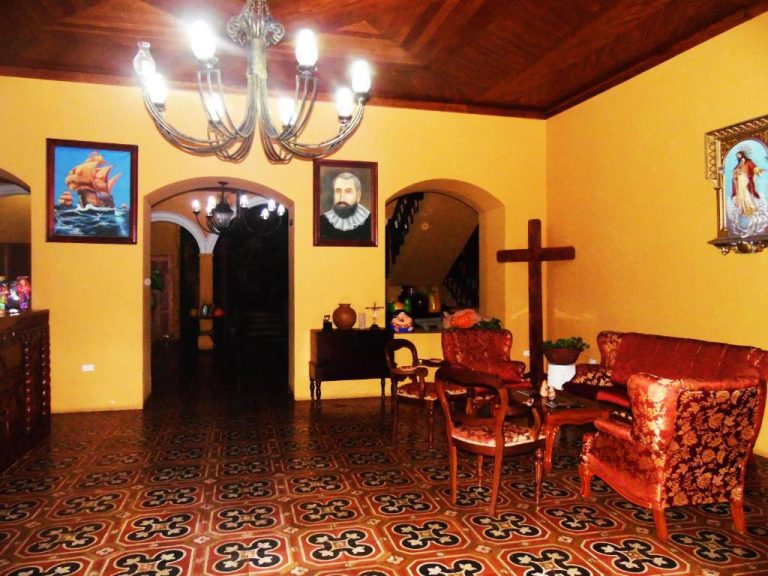 Sala colonial del Hotel Córdoba con crucifijo, sillones rojos, araña de luces y pinturas religiosas en paredes amarillas