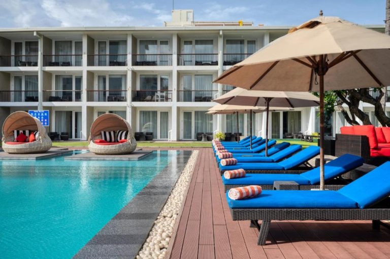 Piscina exterior con tumbonas azules y sombrillas del Camelot Beach Hotel frente al edificio principal