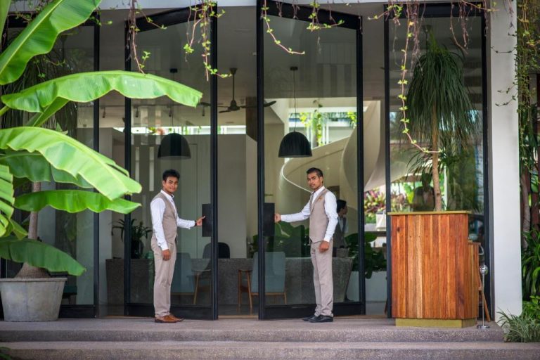 Dos recepcionistas abriendo las puertas de cristal de la entrada principal del Viroth's Hotel rodeada de vegetación tropical
