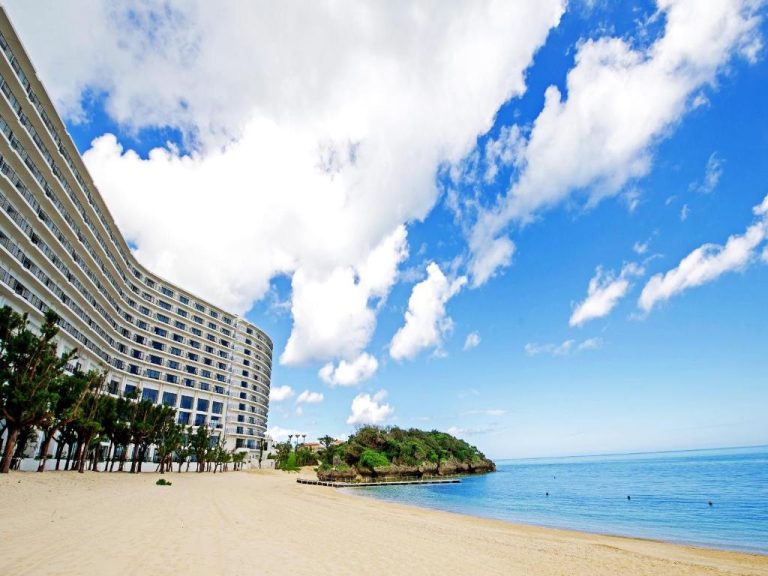 Fachada curva del Hotel Monterey Okinawa Spa & Resort junto a la playa de arena blanca y mar azul