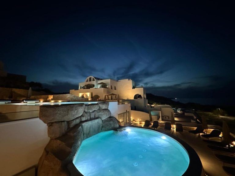 Piscina exterior iluminada de noche con arquitectura cicládica del hotel Aelia Thalassa