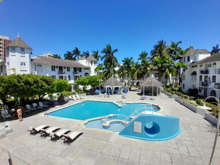 Piscina exterior con tobogán y tumbonas del Beach Studio 21 rodeada de palmeras y edificios blancos