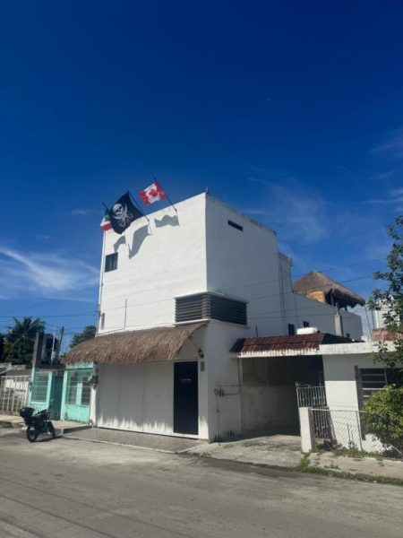 Fachada exterior del La Perle Noire Dive Hotel Cozumel con banderas en la azotea y palapa en la entrada