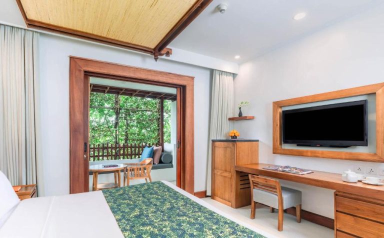 Habitación deluxe con escritorio, TV y balcón privado rodeado de vegetación en Legian Beach Hotel