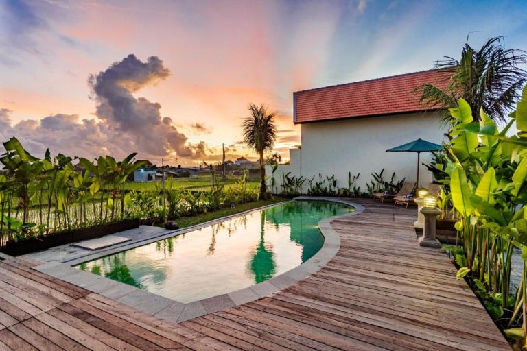Piscina exterior con terraza de madera y vistas al arrozal al atardecer en Svadarma Bali Villa