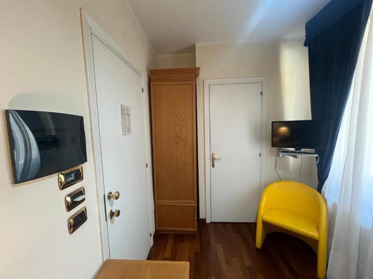 Entrada de habitación del Hotel Liberty con armario de madera, silla amarilla y televisores en la pared