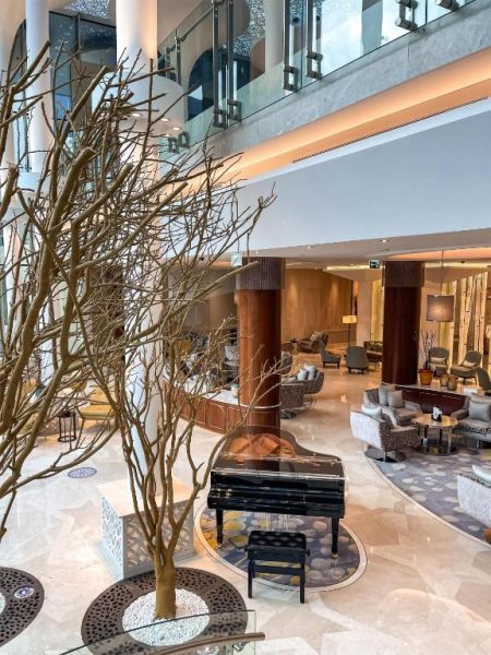 Elegante lobby del Kempinski Hotel Muscat con piano de cola negro y árboles decorativos en atrio de doble altura