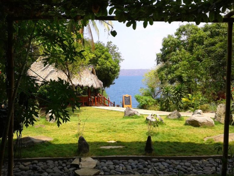 Jardín tropical con palapa frente al lago en Laguna Beach Club, rodeado de vegetación exuberante y rocas naturales