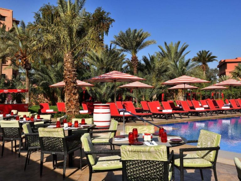 Terraza con mesas y piscina exterior del Mövenpick Hotel Mansour Eddahbi Marrakech rodeada de palmeras