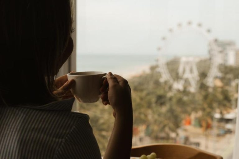 Mujer tomando café con vista al mar y noria desde la habitación del Venti Hotel By Brisa Hotéis
