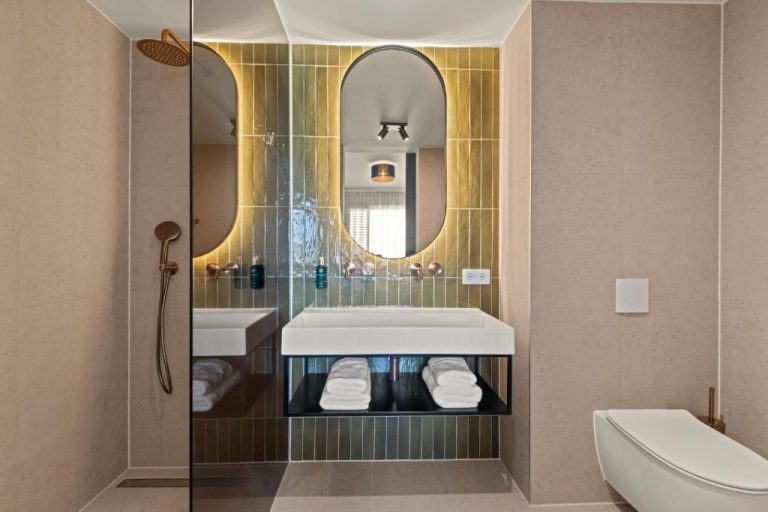 Baño de lujo con azulejos dorados, espejo ovalado iluminado y ducha en The Ivy Blue Bay Curaçao