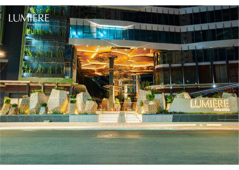 Fachada iluminada de noche con entrada principal del Lumière Riverside, decorada con rocas ornamentales y techo dorado