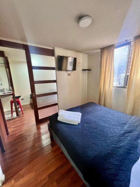 Habitación doble con cama azul y TV en Dptos Merced Santiago, suelo de madera y ventana