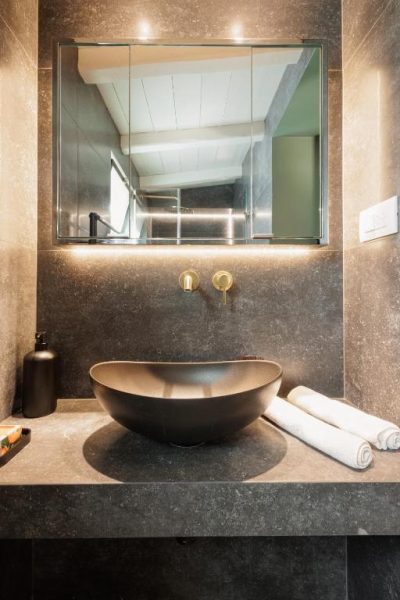 Baño moderno con lavabo negro sobre encimera de piedra y grifería dorada en Hotel Aritis 3