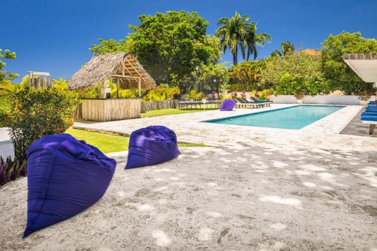 Piscina exterior con palapa y puffs morados en El Encuentro Surf Lodge rodeada de palmeras