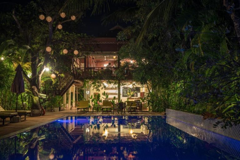 Piscina exterior iluminada de noche rodeada de vegetación tropical en Sambor Village Hotel