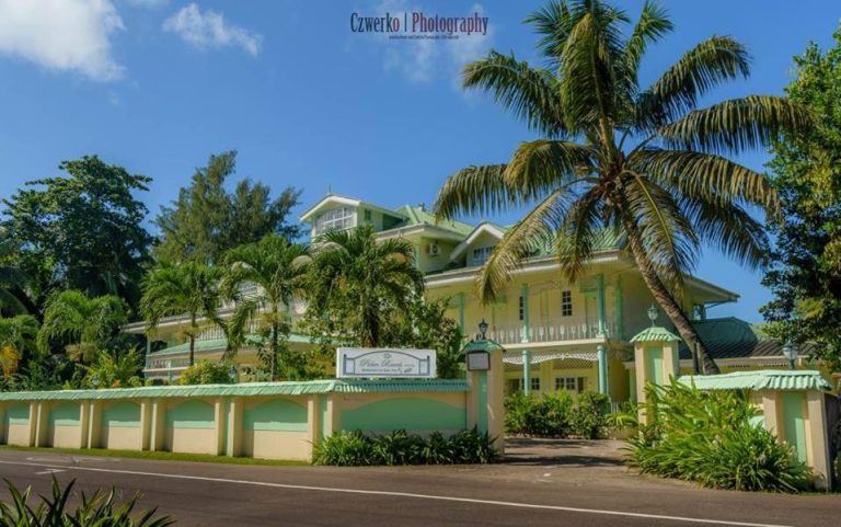 Fachada exterior del Palm Beach Hotel rodeada de palmeras tropicales y jardines exuberantes bajo cielo azul